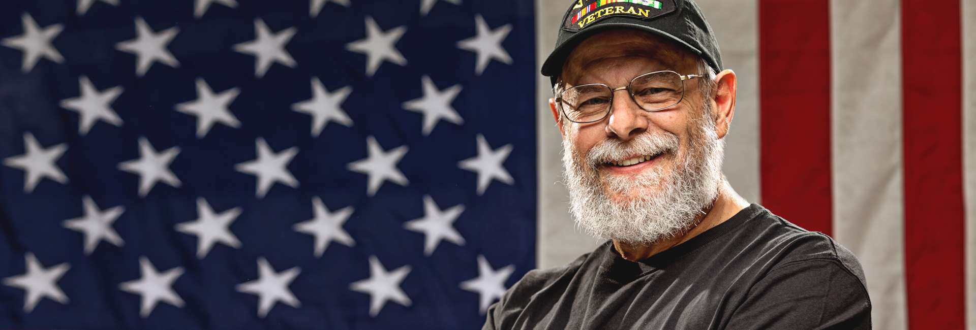 Vietnam Veteran with a beard standing in front of an American flag.